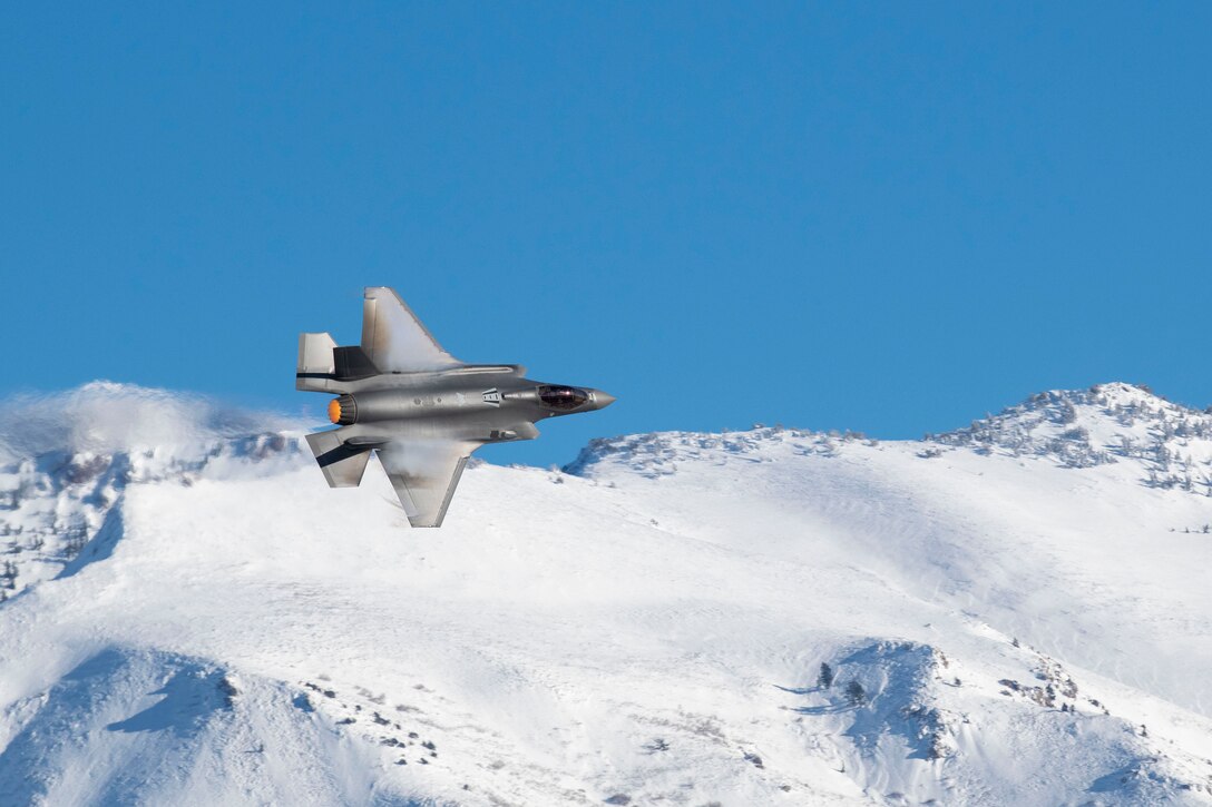 A fighter jet flies near a snow covered mountain range.