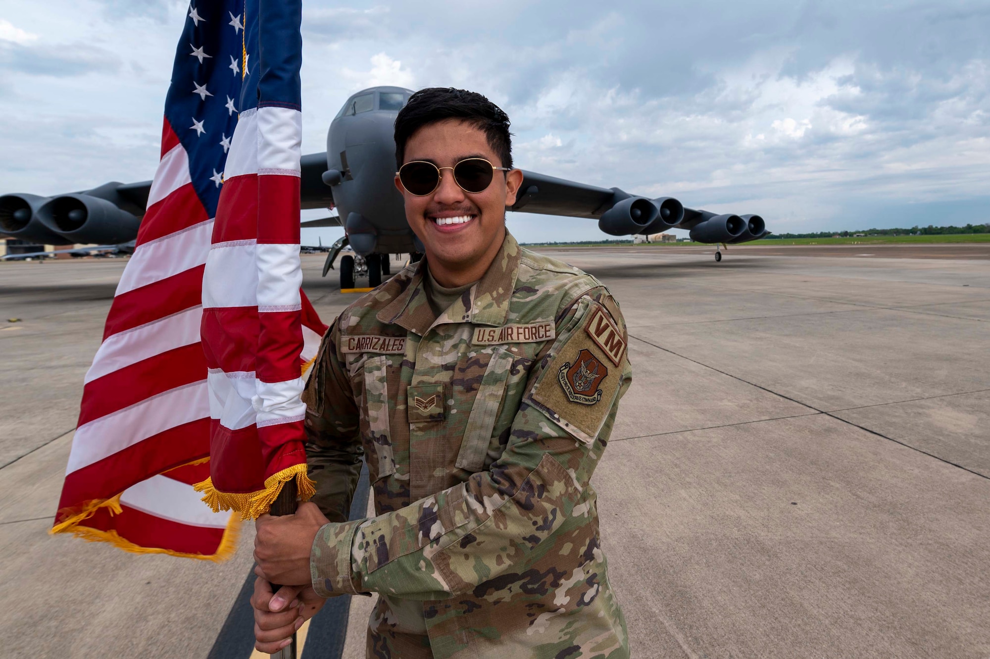 One Airman stands on the flight line, in front of a B-52, holding an American flag. He has on aviator sunglasses and a big smile.