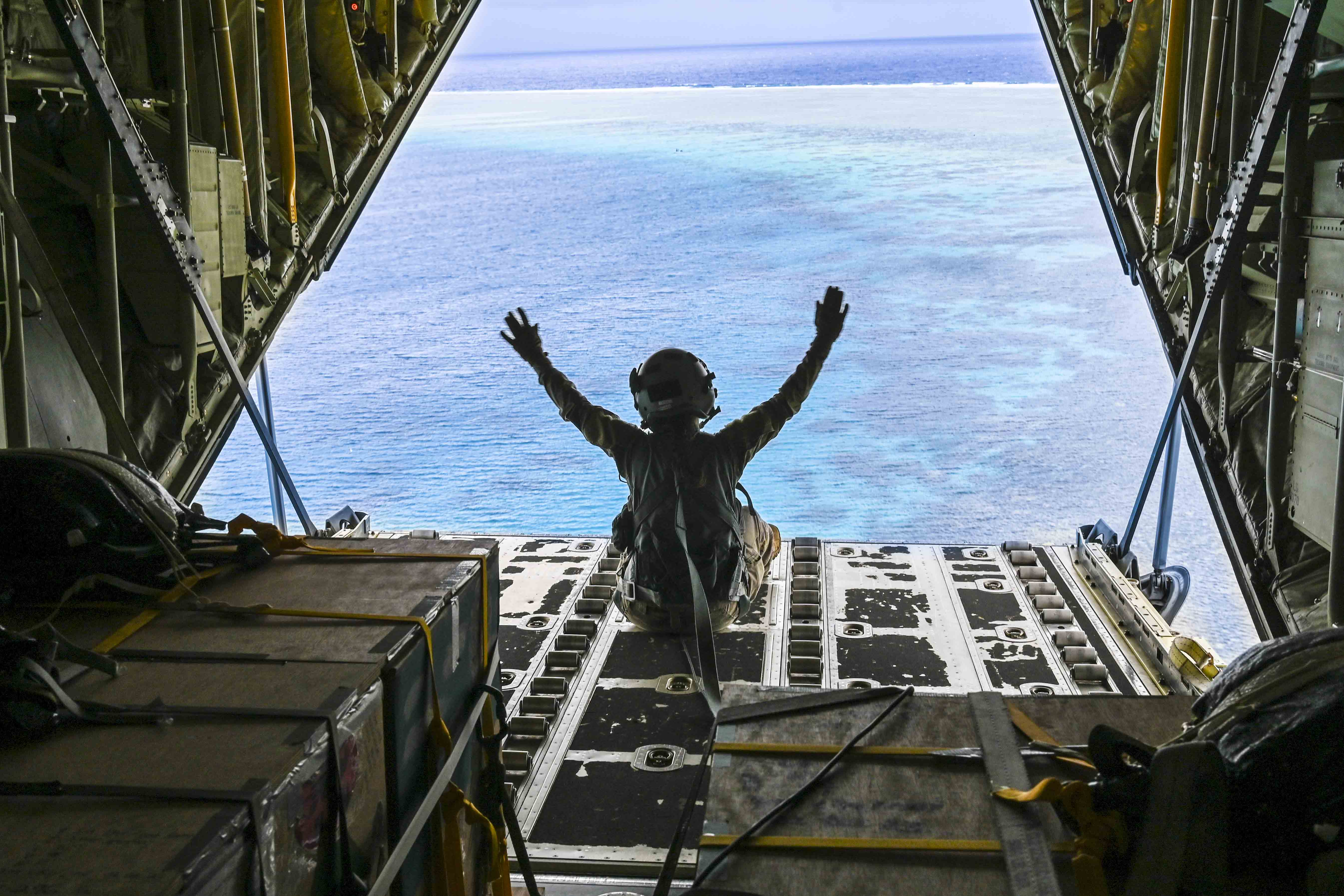 An airman, shown from behind, sits with both hands raised in an open aircraft flying over water.