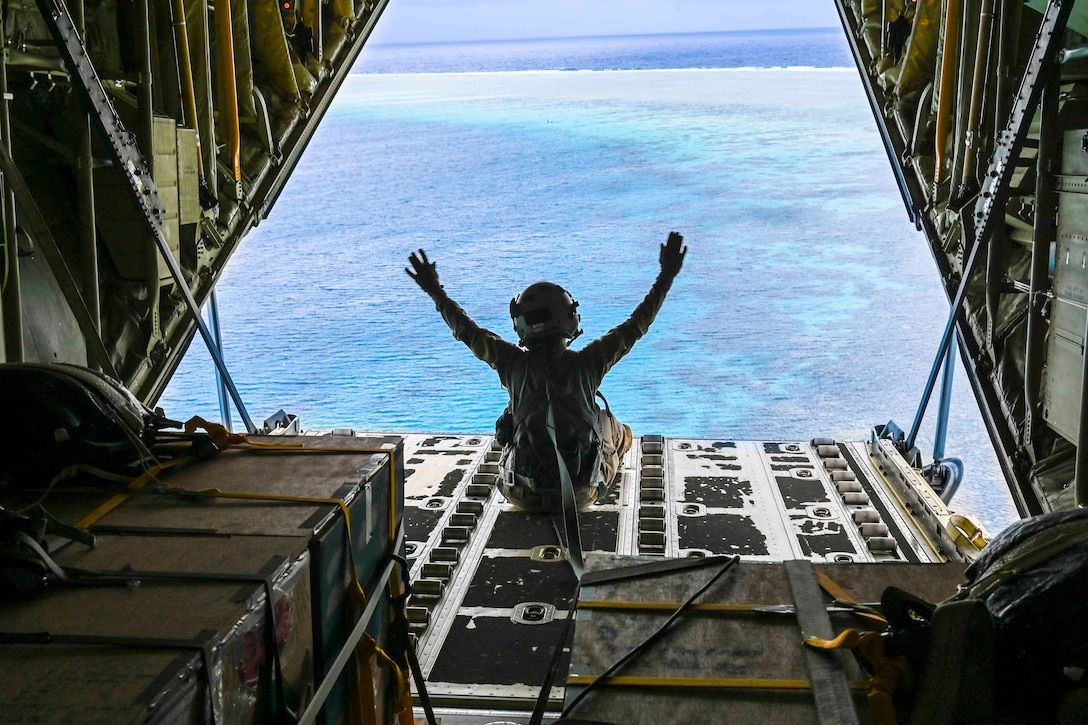 An airman lifts their hands above their head while sitting at the back of an airborne aircraft above a body of water.