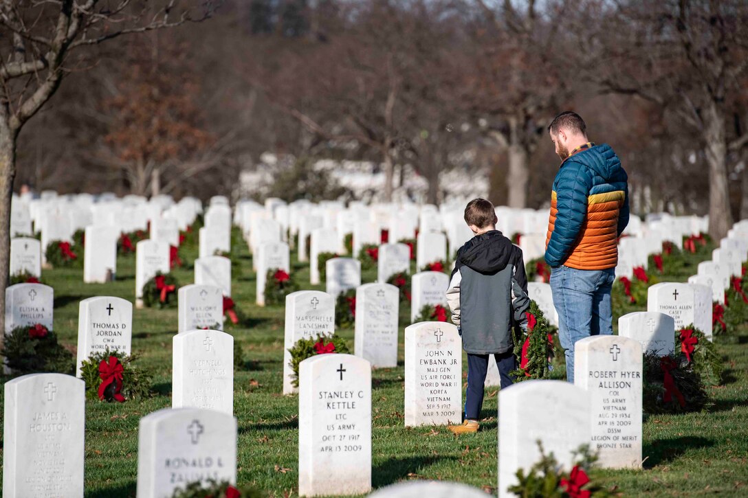 An adult and a child look at a headstone in a cemetery while the child holds onto a wreath.