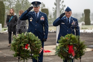 Two uniformed Air Force service members salute in front of ceremonial wreaths
