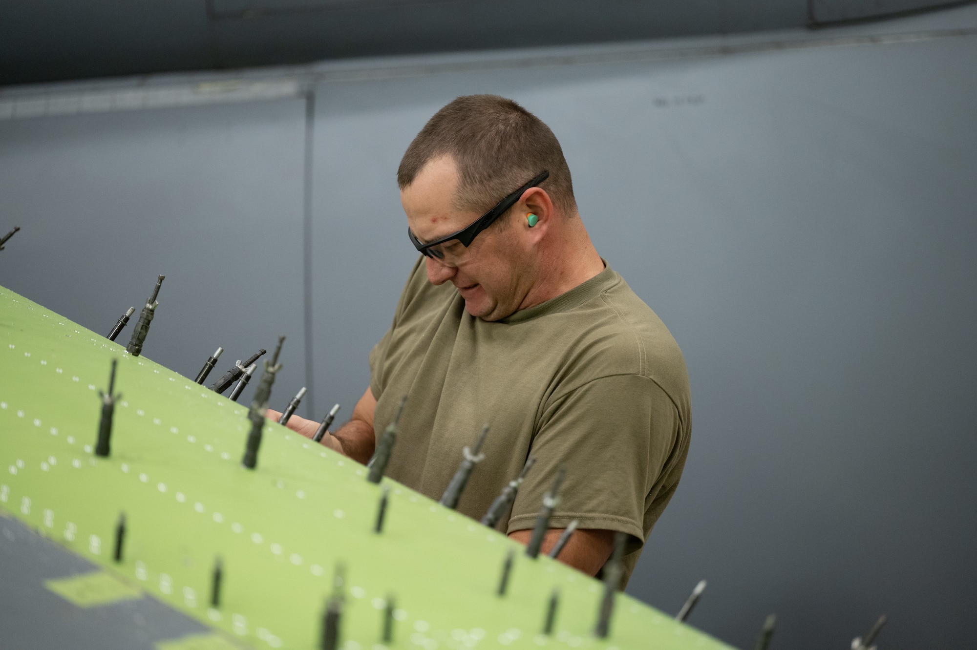 Master Sgt. Jacob Harrington, 434th Maintenance Squadron aircraft structural maintenance journeyman, works on the substructure of a KC-135R Stratotanker’s wing. The foreground shows the wing's substructure, a bright green, and Harrington in the background, conducting the repair with a tool. He is wearing safety glasses and ear plugs.