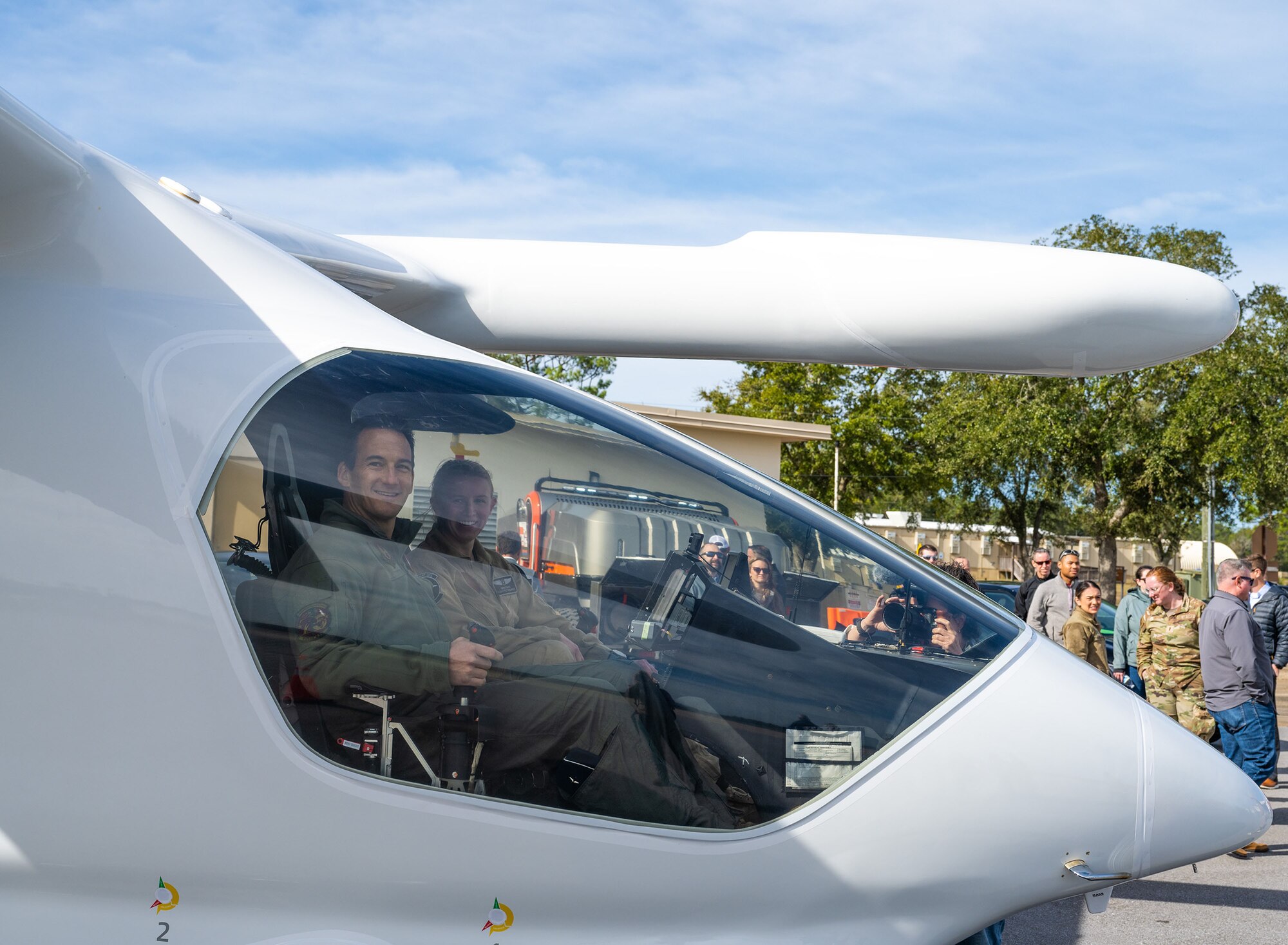 Two military members sit in the cockpit of an electric aircraft and look out and smile at the viewers.