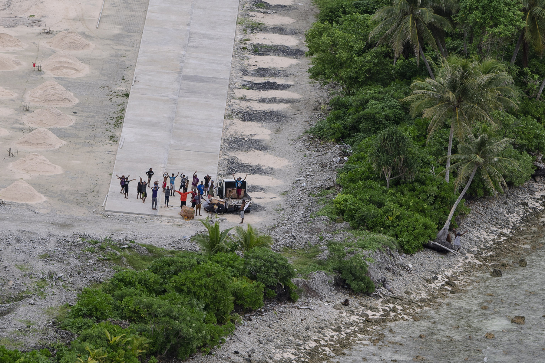 About 20 people, shown from above, stand and wave at the end of a rough airstrip on an island.