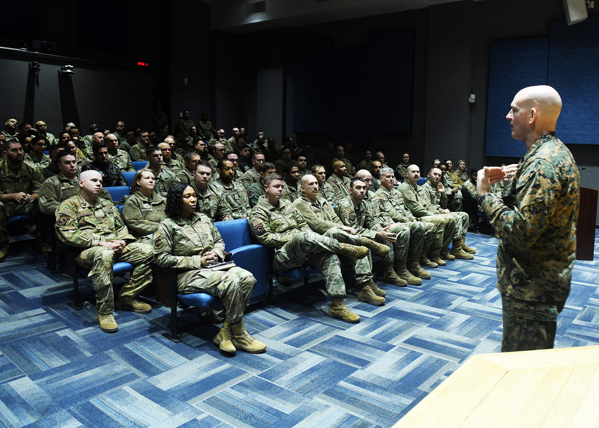 military personnel fill an auditorium listening to a speaker.