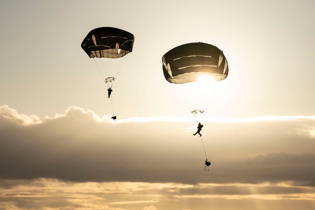 Two soldiers wearing parachutes descend over Alaska. Sun rays can be seen peeking through clouds in the background.