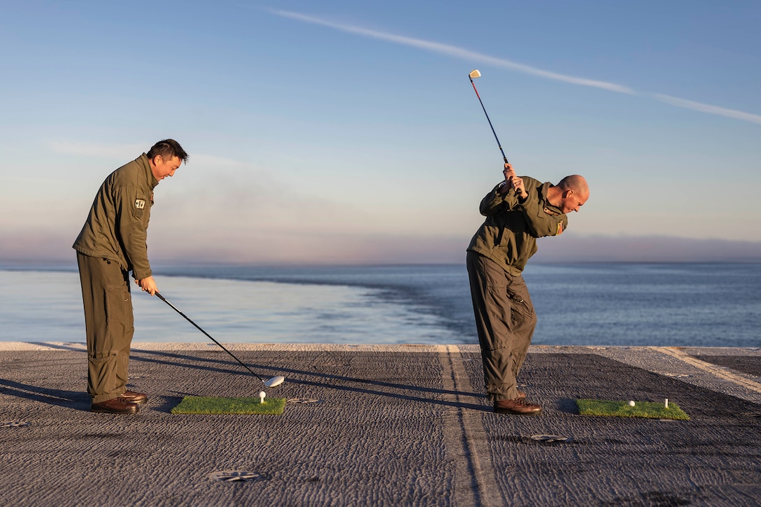 Two sailors prepare to hit golf balls off the flight deck of a Navy ship at sea.