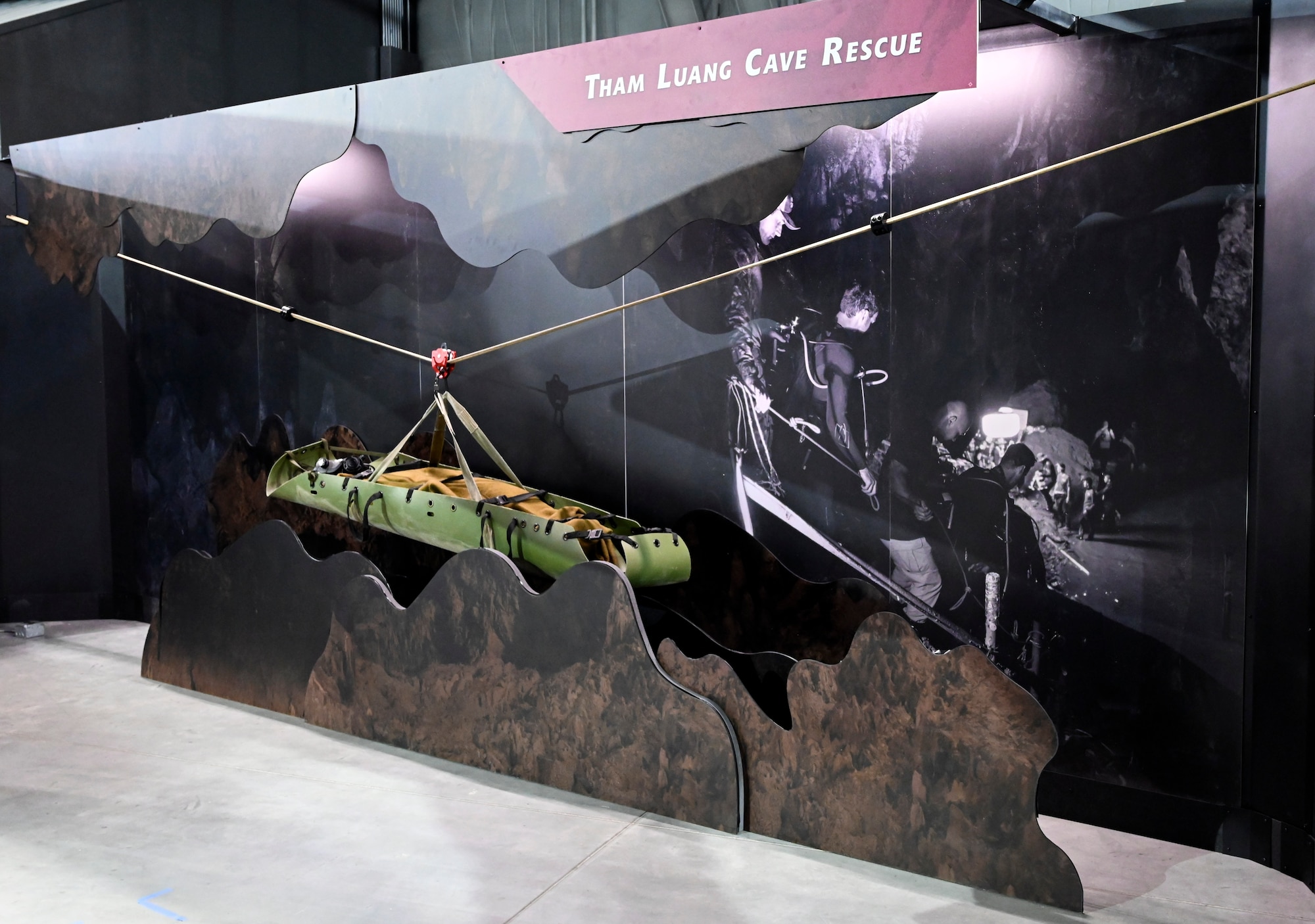 Image of a sked pararescue stretcher in the humanitarian exhibit's Rescue mission. The stretcher is olive drab green and is suspended from a horizonal cable to simulate how they were able to safely pull the kids out of the Thai caves.