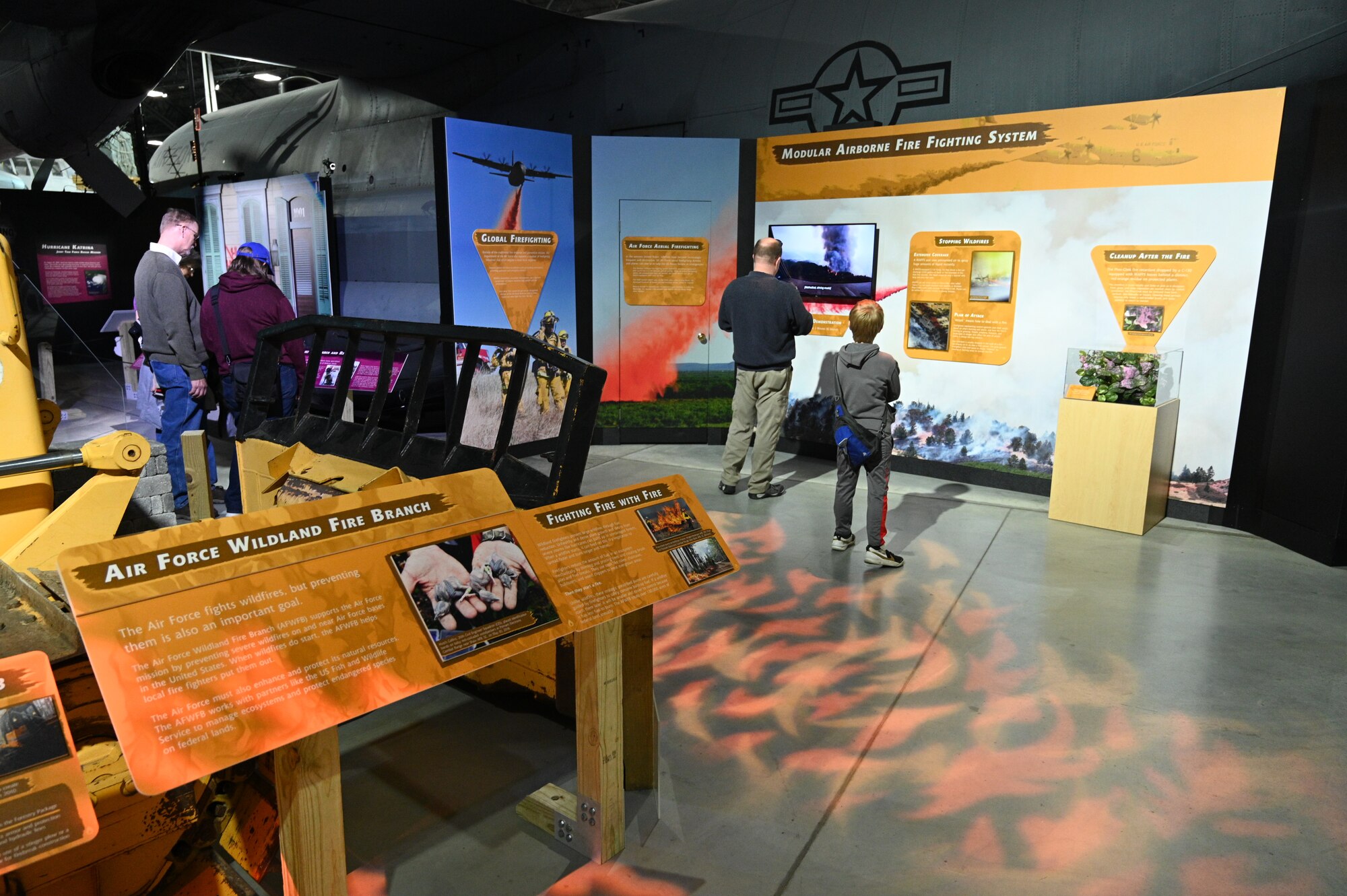 Image of the Global Firefighting mission portion of the Humanitarian Exhibit. On the left is a yellow panel with text descriptions in the exhibit. In the front of the picture is a family looking at a tall panel describing the wildlife firefighting process.