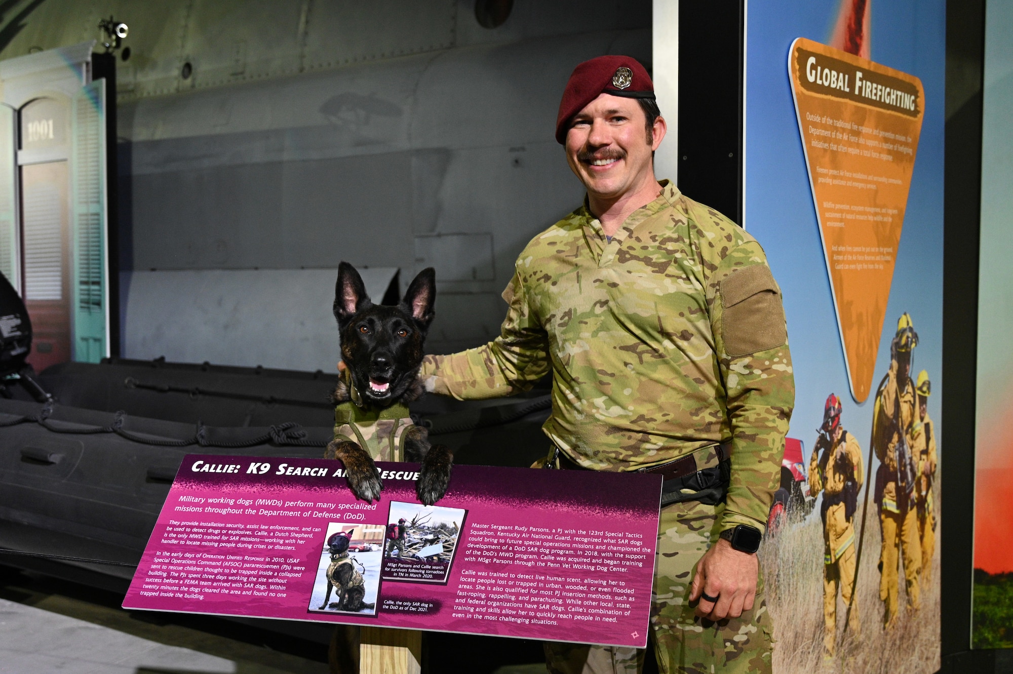 Callie, a Dutch Shepherd military working dog stands in front of the display honoring his service in the Humanitarian exhibit, with his handler Master Sgt. Rudy Parsons.