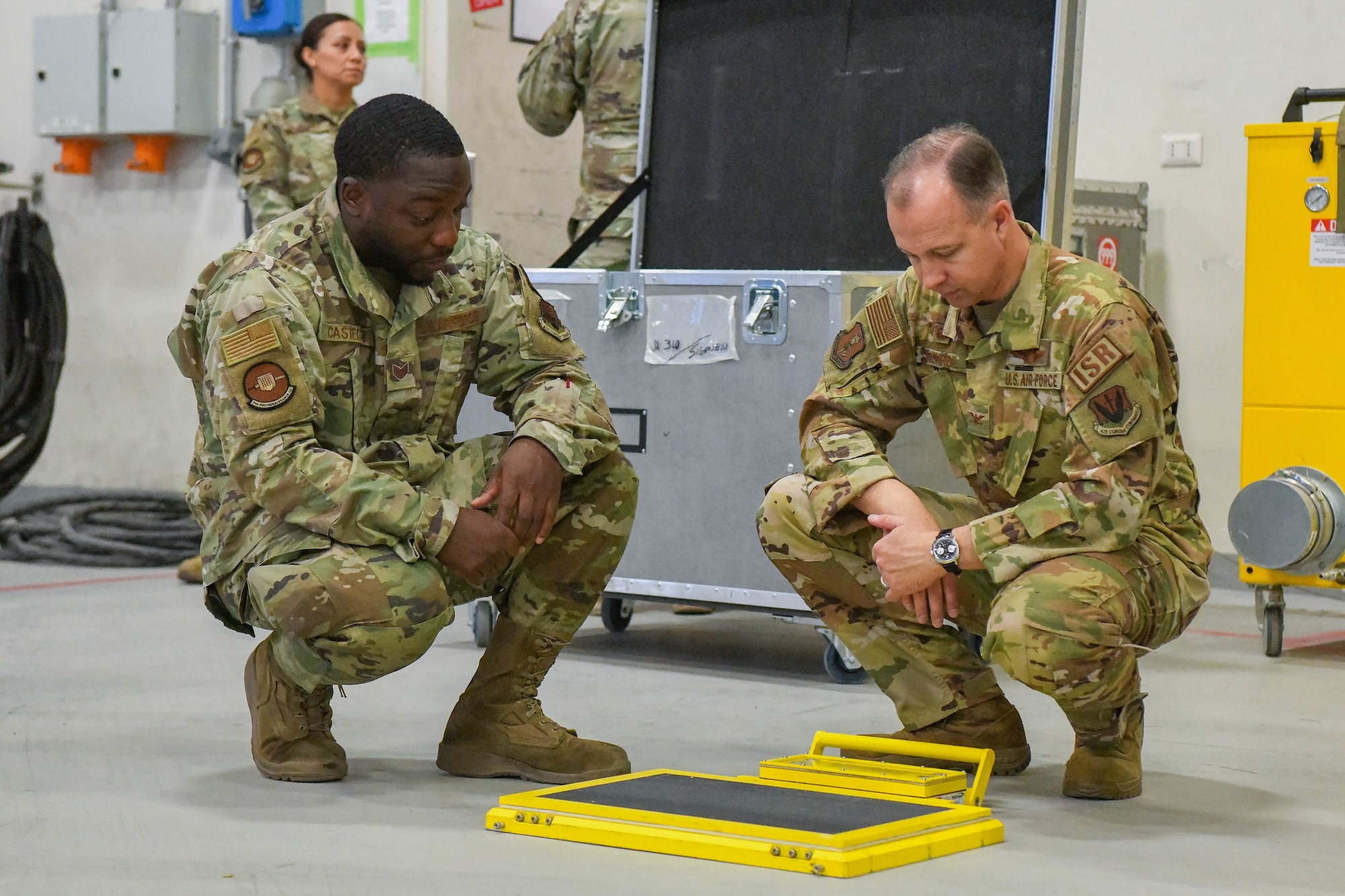 Two men in a military uniform squat down while looking at an industrial scale.