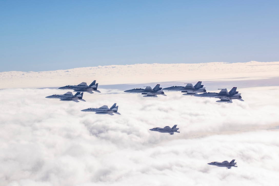 Eight military aircraft fly over clouds during daylight.