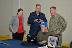 One man with a blue baseball cap sits at a laptop working while a woman and two men, one in a Navy uniform watch the seated man work.