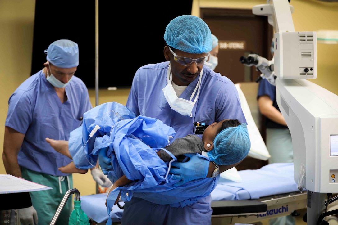 A sailor dressed in medical protection equipment carries a patient in an operating room.