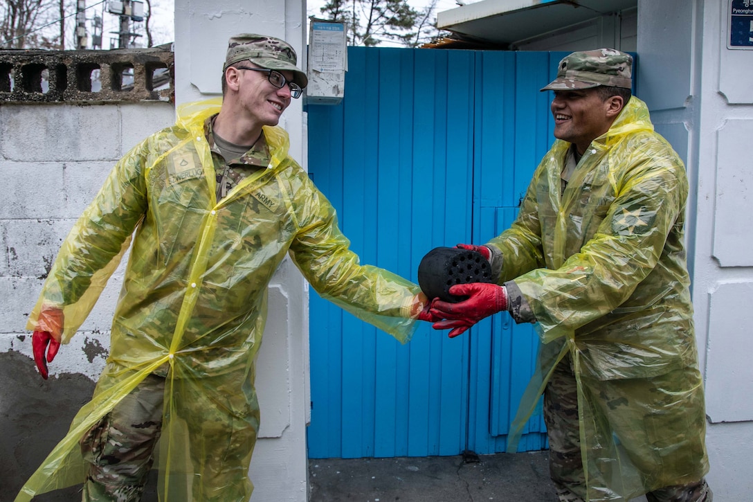 A smiling, uniformed soldier wearing a plastic raincoat and gloves hands coal briquettes to another soldier dressed in the same gear.