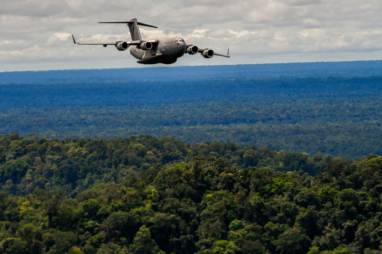 A military aircraft flies over a wooded area during daylight.