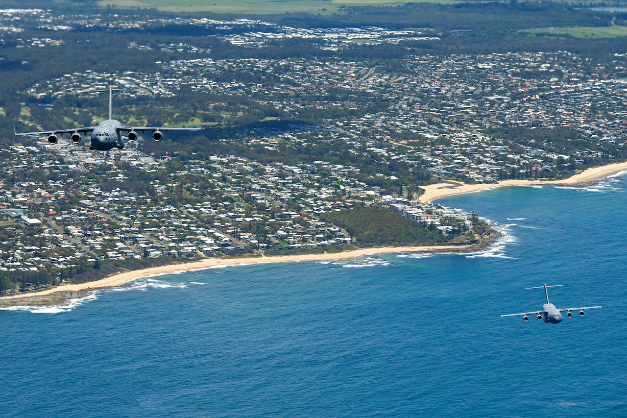 Military aircraft fly over blue water during daylight. A shoreline can be seen in the background.