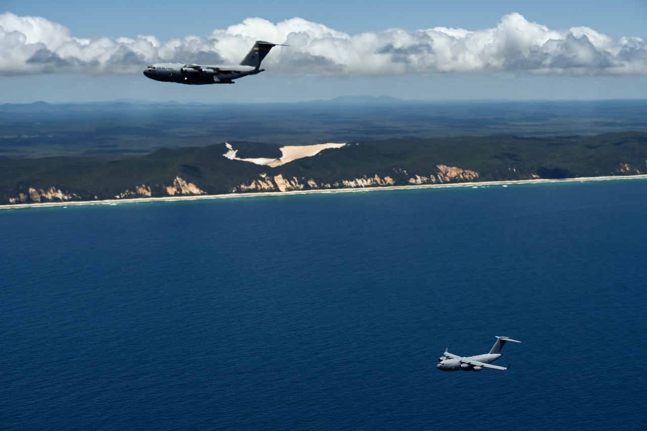 Military aircraft fly over blue water during daylight. A shoreline can be seen in the background.