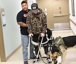 Saron Om, a physical therapist at Womack Army Medical Center giving Sgt. William Keelan, accident victim assigned to the Soldier Recovery Unit Fort Liberty cues to improve walking movement patterns and assist with recovery from any loss of balance as Koda his service dog stands by.