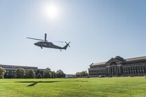 a UH-60 Blackhawk helicopter is hovering, about to land, on a green lawn near the National Defense University. There is a large red brick building with a dome on the right side of the picture in the distance.