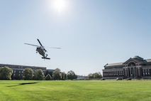 a UH-60 Blackhawk helicopter is hovering, about to land, on a green lawn near the National Defense University. There is a large red brick building with a dome in the background on the right of the picture.