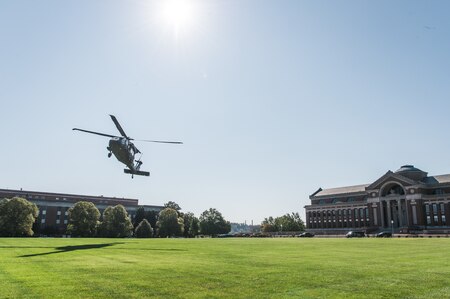 a UH-60 Blackhawk helicopter is hovering, about to land, on a green lawn near the National Defense University. There is a large red brick building with a dome in the background on the right of the picture.