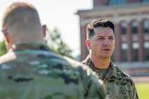 Two Army soldiers in fatigues are waiting to load into a UH-60 Blackhawk helicopter, which isn't in view. There is a large red brick building in the background