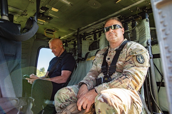 An Army soldier in fatigues and wearing sunglasses is sitting inside a UH-60 Blackhawk helicopter with a civilian who is wearing dark cloths.