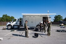 Several Army service members and members of the FBI are standing around a metal frame as they assemble it in front of a white trailer.