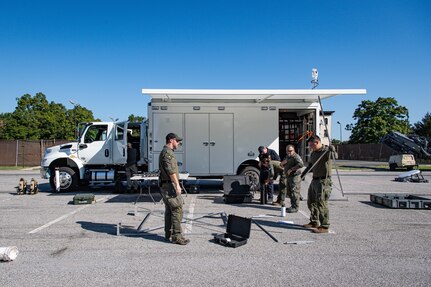 Several Army service members and members of the FBI are standing around a metal frame as they assemble it in front of a white trailer.