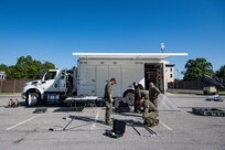Several Army service members and members of the FBI are standing around a metal frame as they assemble it in front of a white trailer.