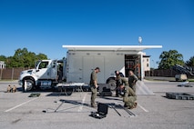 Several Army service members and members of the FBI are standing around a metal frame as they assemble it in front of a white trailer.