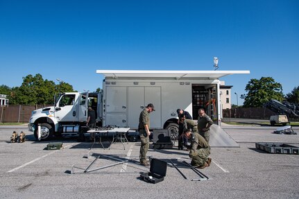 Several Army service members and members of the FBI are standing around a metal frame as they assemble it in front of a white trailer.