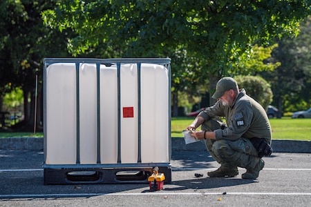 A member of the FBI is squatting near a large white plastic container with a piece of paper in his hand. He is wearing all green utility clothes.