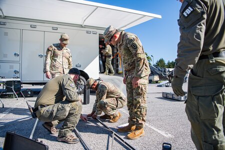 Several Army service members and members of the FBI are standing around a metal frame as they assemble it in front of a white trailer.