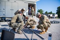 Several Army service members and members of the FBI are standing around a metal frame as they assemble it in front of a white trailer.