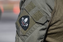 A closeup of a black shoulder patch that is attached with velcro to the shoulder of someone wearing a dark green military utility uniform.