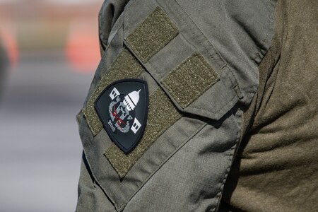 A closeup of a black shoulder patch that is attached with velcro to the shoulder of someone wearing a dark green military utility uniform.