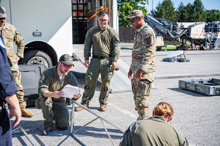 Several Army service members and members of the FBI are standing around a metal frame as they assemble it in front of a white trailer.