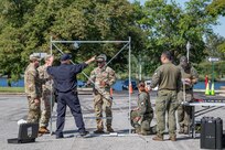 Several service members in Army fatigues are standing around a tall metal frame with another member in a dark blue work outfit pointing to something on the frame. It is setup in a parking lot with a few small dark cases on the ground around it.