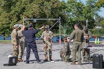 Several service members in Army fatigues are standing around a tall metal frame with another member in a dark blue work outfit pointing to something on the frame. It is setup in a parking lot with a few small dark cases on the ground around it.