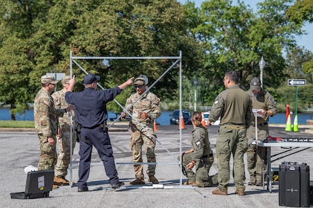 Several service members in Army fatigues are standing around a tall metal frame with another member in a dark blue work outfit pointing to something on the frame. It is setup in a parking lot with a few small dark cases on the ground around it.