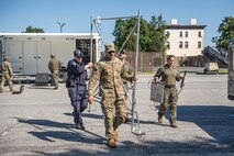 Service members in Army fatigues and other members in all dark blue or green working outfits are carrying a tall metal frame across a parking lot. In the background is a white trailer with a few dark green cases out on the ground in front of it.