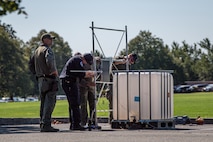 Four Service members from 55th Explosive Ordinance Disposal (EOD) Company and EOD agents from the FBI’s Washington Field Office are standing near a metal frame, which is near a large square-shaped plastic container.