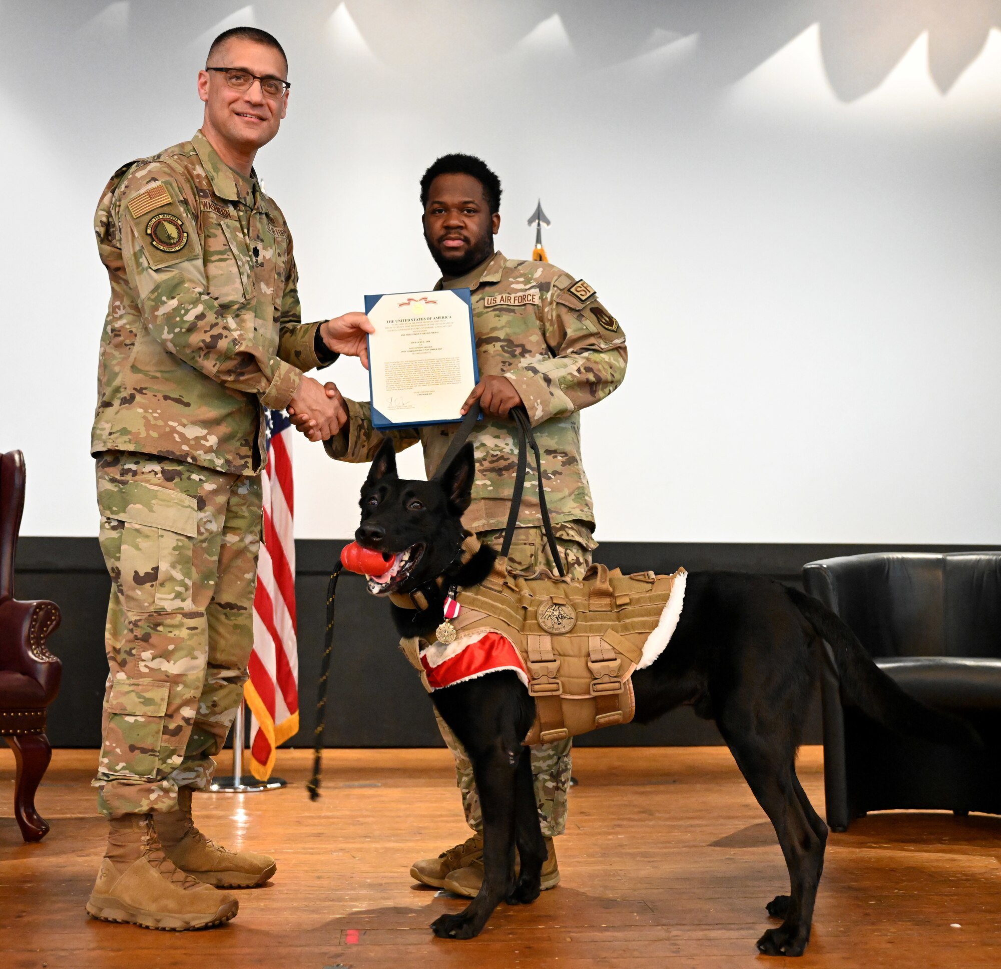 U.S. Air Force Lt. Col. Ben Washburn, left, 48th Security Forces Squadron commander, presents the Meritorious Service Medal and certificate to Military Working Dog Cchuy, at a double-retirement ceremony with MWD Ukkie, at Royal Air Force Lakenheath, England, Dec. 13, 2023. Cchuy is being adopted by his final handler, Staff Sgt. Ben Kaupp (not pictured) and his family, but stood on the stage with another handler, due to the tradition of officially handing over the retired MWD to his or her new family. (U.S, Air Force photo by Karen Abeyasekere)
