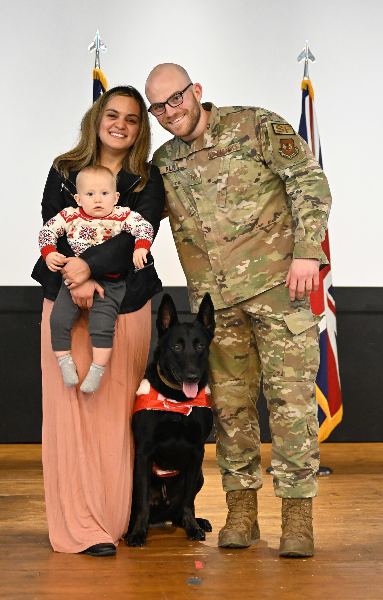 U.S. Air Force Staff Sgt. Ben Kaupp, right, and his wife and son get their first family photo taken with newly retired Military Working Dog Cchuy, who they have adopted, at the end of the K9’s official retirement ceremony at Royal Air Force Lakenheath, England, Dec. 10, 2023. Cchuy amassed more than 5,900 in all-weather specialized explosive search, deterrence and intruder detection operations, to provide a safe and secure environment for the mission accomplishment of the 48th Fighter Wing. Kaupp was Cchuy’s final handler and formed a special bond with the Belgian Malinois, and as soon as he knew the MWD was retiring, he and his wife knew they wanted Cchuy to become part of their family. (U.S. Air Force photo by Karen Abeyasekere)