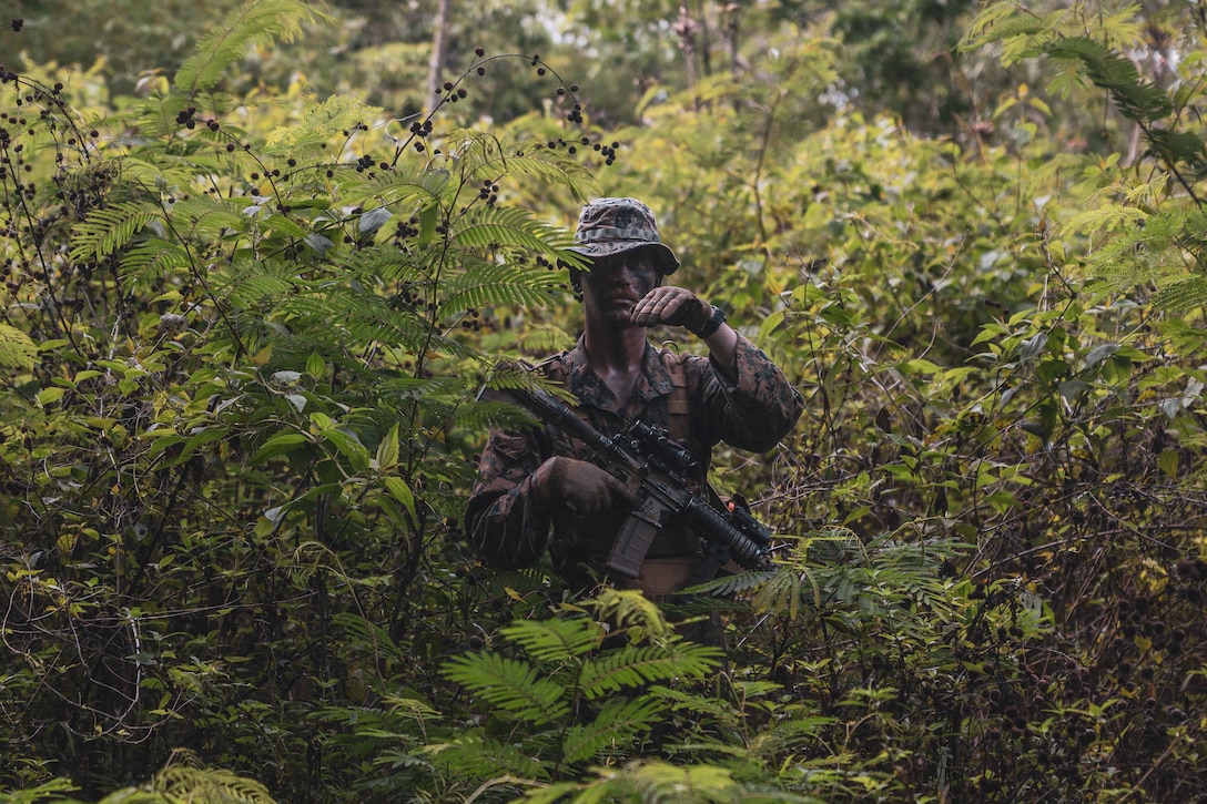 A Marine stands in some bushes and uses hand signals to communicate.