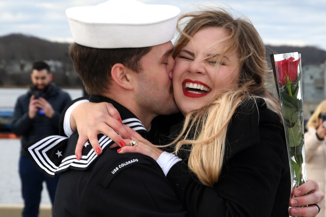 A sailor hugs his wife as she laughs.