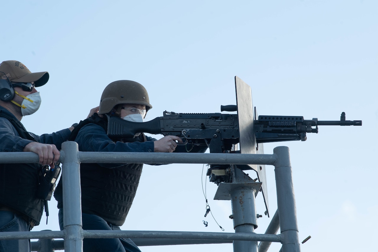 A service member fires a weapon while another service member, wearing hearing protection, looks on.